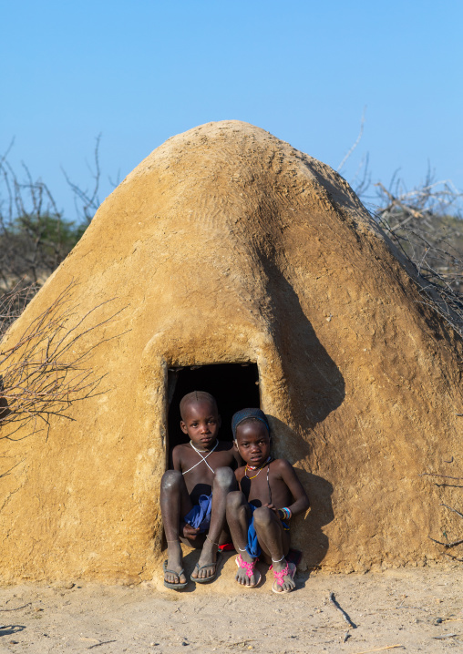 Mucubal tribe children sitting at the entrance of their hut covered of adobe, Namibe Province, Virei, Angola