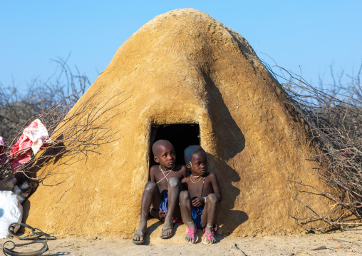 Mucubal tribe children sitting at the entrance of their hut covered of adobe, Namibe Province, Virei, Angola