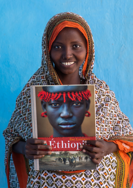 Portrait of a veiled smiling afar tribe girl with sharpened teeth holding eric lafforgue book, Afar Region, Afambo, Ethiopia