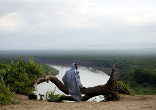 Karo Tribe Man In The Morning Sitting On A Tree Trunk Over The Omo River, Korcho Village, Omo Valley, Ethiopia