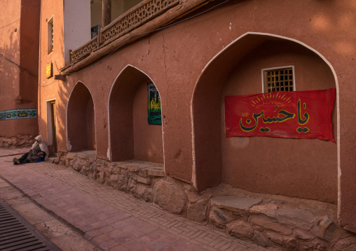 Woman Sitting In Front Of An Ancient Building With Ashura Decoration In Zoroastrian Village, Isfahan Province, Abyaneh, Iran