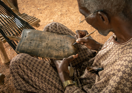 Senior man sit on a hammock writing koran on a wood board, Savanes district, Kouto, Ivory Coast