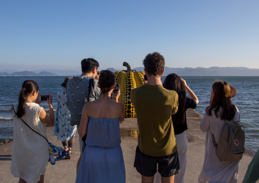 Tourists taking pictures of the yellow pumpkin by Yayoi Kusama on pier at sea, Seto Inland Sea, Naoshima, Japan