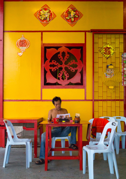 Mature Man Reading A Book In Front Of His Yellow House In The Weld Quay Clan Jetty, Penang Island, George Town, Malaysia