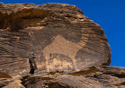 Tourist standing in front of a life-sized camel petroglyph on a rock, Najran Province, Thar, Saudi Arabia