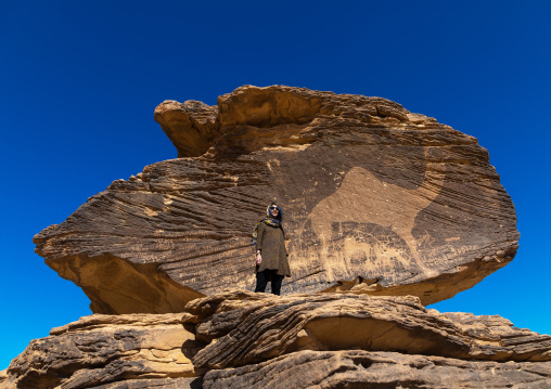 Tourist standing in front of a life-sized camel petroglyph on a rock, Najran Province, Thar, Saudi Arabia